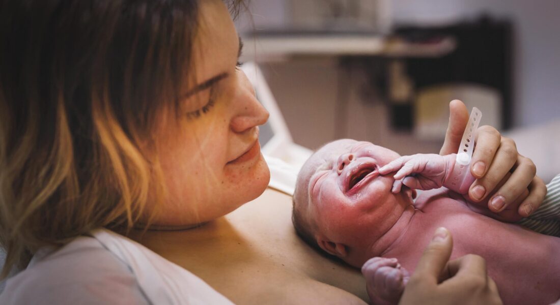 På bilden ser vi en nybliven mamma vars ansiktsuttryck av lättnad och lycka speglar den djupa emotionella resan. Hon ser ömt på sitt barn, vars första skrik och livliga uttryck symboliserar början på ett nytt liv. Barnmorskan, vars händer varsamt stöder bebisen, förbereder en första medicinsk undersökning som säkerställer att allt står rätt till.