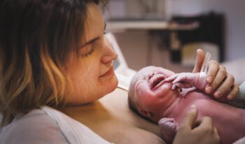 På bilden ser vi en nybliven mamma vars ansiktsuttryck av lättnad och lycka speglar den djupa emotionella resan. Hon ser ömt på sitt barn, vars första skrik och livliga uttryck symboliserar början på ett nytt liv. Barnmorskan, vars händer varsamt stöder bebisen, förbereder en första medicinsk undersökning som säkerställer att allt står rätt till.